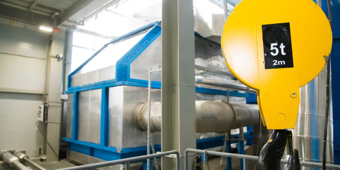 Digital crane scale suspended between overhead crane hook and heavy steel load in an industrial manufacturing facility