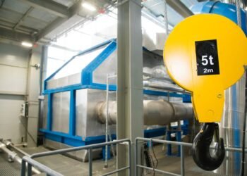 Digital crane scale suspended between overhead crane hook and heavy steel load in an industrial manufacturing facility