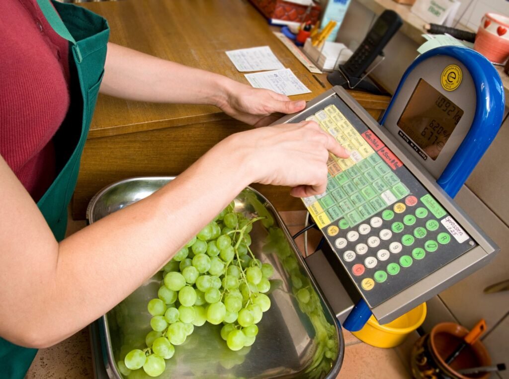 Produce department associate using a price computing scale with PLU codes to weigh and label fresh vegetables with a price barcode before they reach the checkout lane