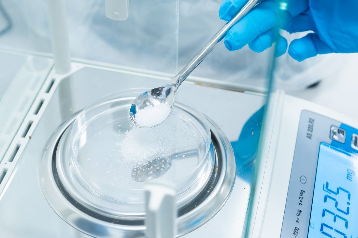Gloved hand using a spatula to add white granules to a glass weighing dish inside a balance draft shield